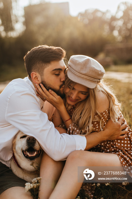 Guy in white shirt hugs and kisses his beloved girlfriend on cheek. Woman in polka-dot dress and her husband fooling around with their dog in park