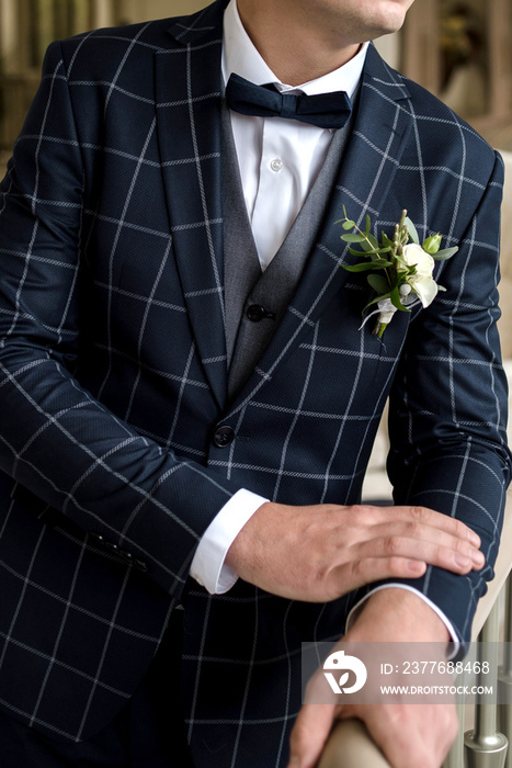 Groom in white shirt, plaid suit and bow tie with buttonhole waiting for bride indoors, copy space. Morning of the groom. Wedding concept