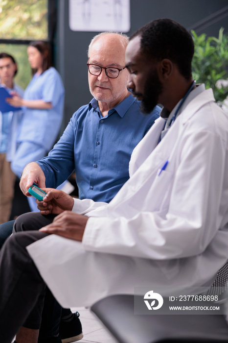 African american medic consulting senior patient with glucometer instrument while measuring insulin and glucose level from blood sample. Elderly man having checkup visit consultation in hospital lobby