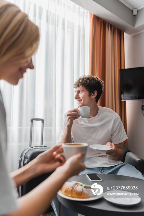 smiling man sitting in hotel room with coffee cup and looking away near girlfriend on blurred foreground.