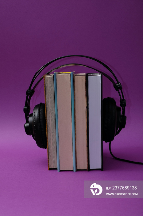 Large black headphones hung from the top on books in an upright position on a purple background. Text space. Audio books, book lovers, podcast, concept. Minimal style.