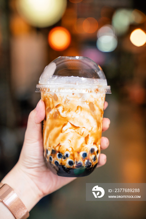 Young girl is holding and showing a cup of brown sugar flavored tapioca pearl bubble milk tea in night market of Taiwan background, close up, bokeh