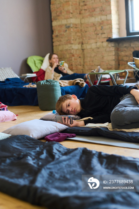 Schoolboy with mobile phone resting on sleeping place on the floor and texting against young female refugee lying on couchette