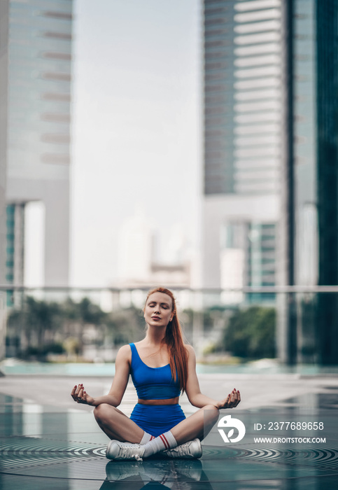 Slim yoga woman in sportswear shorts and top sits in lotus position, asanas with crossed legs on city street in Dubai with eyes closed, feeling pacification, meditating