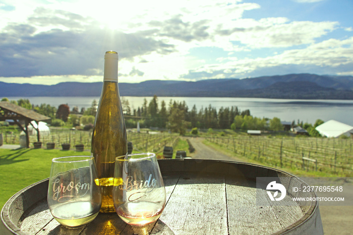 Wedding in the winery - The view on two wine glasses for Bride and Groom with bottle of white wine placed on wooden barrel with the view on Okanagan lake and Kelowna vineyards in the background.