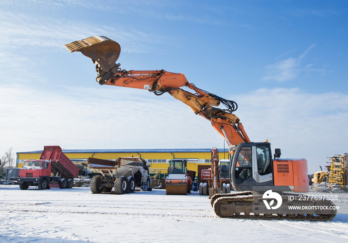 large working machine at the construction site - a crawler excavator. A powerful orange excavator with a raised bucket against the backdrop of a blue sky and other working equipment.