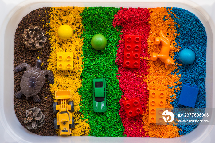 Sensory box with rainbow rice inside. Children’s hands touch the texture and pour.