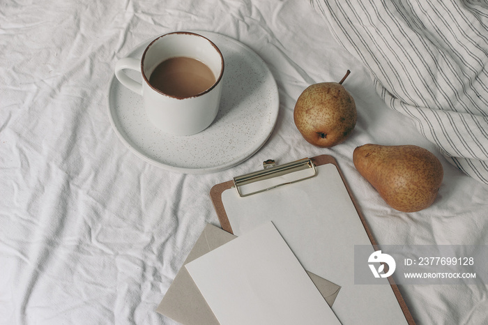 Autumn, summer breakfast in bed composition. Cup of coffee and pear fruit on ceramic plate. Blank greeting card, clip holder mockup White striped linen bedding. Feminine lifestyle composition.