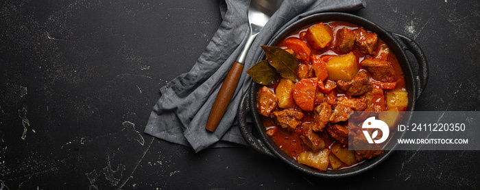 Beef meat stew with potatoes, carrot and delicious gravy in black casserole pot with bay leaves with spoon on black dark rustic concrete background from above, space for text.