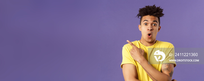 Portrait of surprised, excited young man showing cool new thing, pointing upper left corner, gasping, folding lips, stare camera impressed say wow, standing purple background