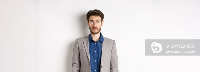 Shocked male entrepreneur in suit drop jaw, staring worried and confused at camera, standing against white background