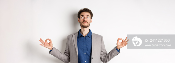 Calm ceo manager in suit relaxing with meditation, looking up and holding hands in zen mudra sign, standing on white background