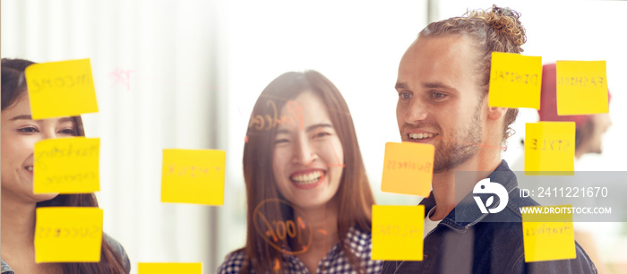 Hipster attractive young caucasian man and young asian chinese girl talk and brainstorming together at glass wall sharing business ideas project plans with post it sticky note. Marketing plan concept.