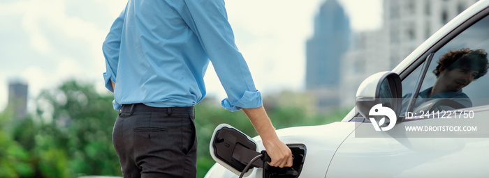 Closeup progressive man holding EV charger plug from public charging station for electric vehicle with background of residential building as concept eco-friendly sustainability energy car concept.