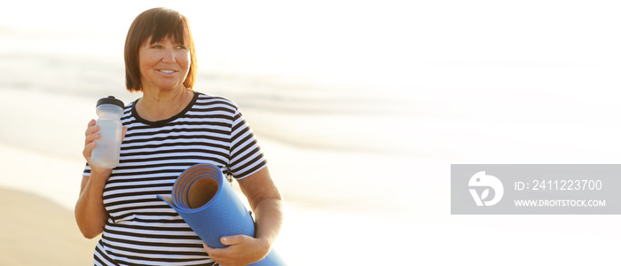 Smiling mature adult woman with bottle of water after exercising outdoors holding yoga mat outdoors on beach by sea on summer. Senior healthy lifestyle, vitality, healthcare concept. copy space