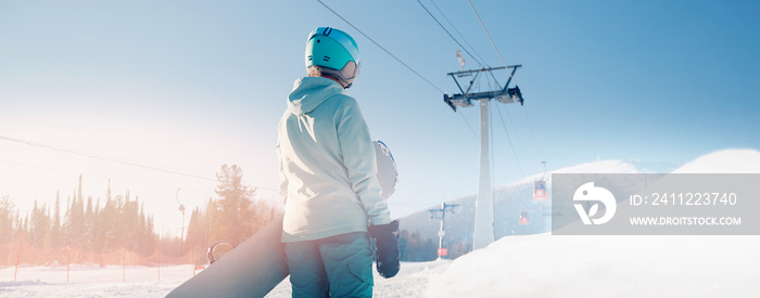 Back young woman snowboarder stands with snowboard sunlight, banner winter ski resort