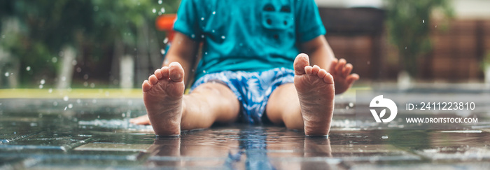 No face photo of a caucasian small boy in blue clothes and barefoot sitting on the ground in water and playing