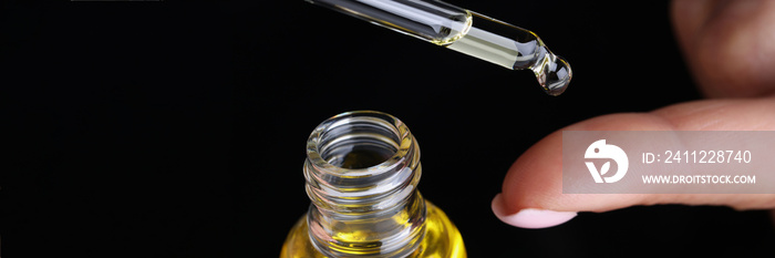 Drop of yellow oil flowing from dropper onto woman finger on black background closeup