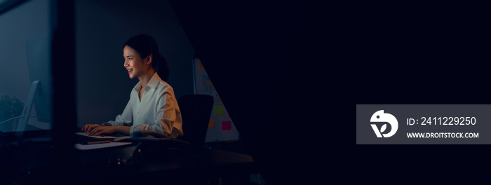 Smiling Asian business woman sitting the table and working on computer in office at night. Take your screen to put on advertising.