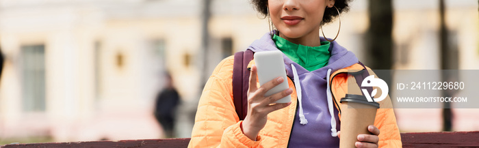 Cropped view of young african american woman with backpack using smartphone and holding coffee to go outdoors, banner