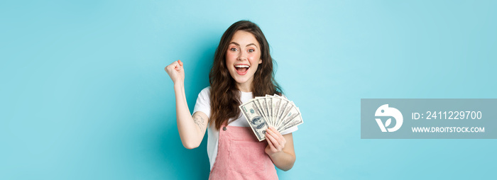 Excited smiling girl fist pump and hold money prize, winning cash, receive income from something, standing happy against blue background