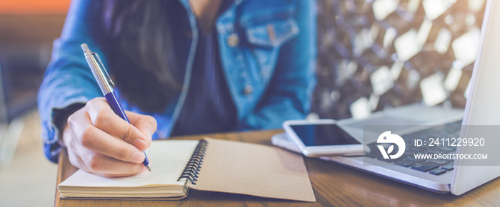 A woman’s hand is writing on a notepad with a pen and a phone with a laptop.Web banner.