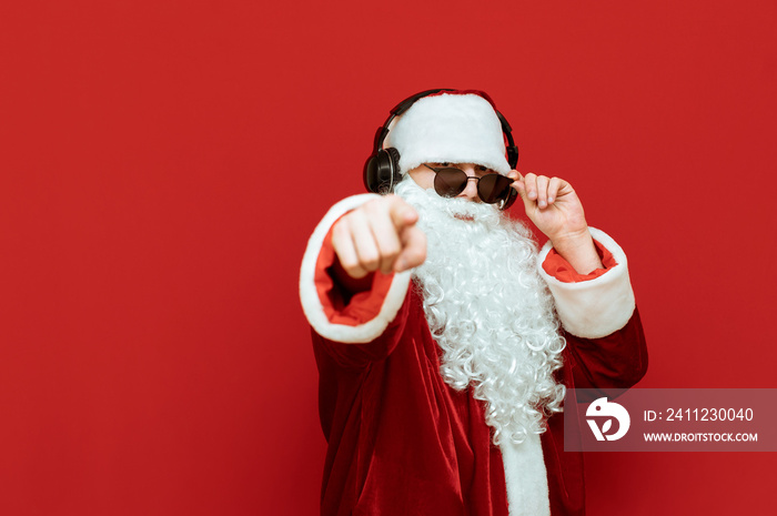 Santa Claus in sunglasses and headphones listens to music, looks into the camera and points his fing