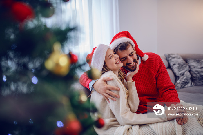 Charming caucasian couple in sweaters and with santa hat on heads sitting on sofa in living room nex