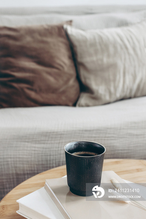 Books, dry bunny tail grass and black cup of coffee on round wooden table. Blurred beige sofa backgr