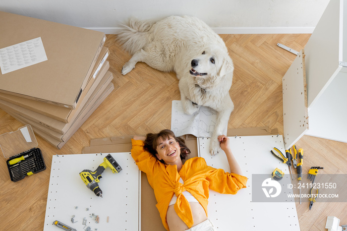 Young happy woman lies on floor with her cute dog, resting while making repairing at new apartment, 