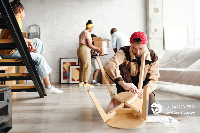 Young furniture assemblage specialist in workwear standing on knees on the floor and assembling new 