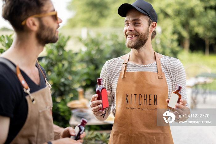 Two handsome male friends in aprons cooking food on a picnic standing together with alcohol drinks a