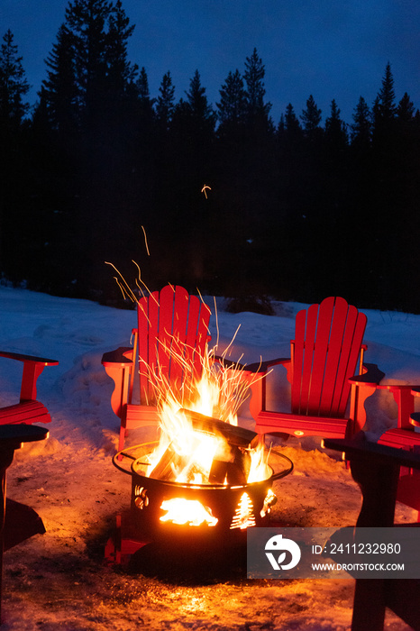 Cozy/romantic evening scene of warming bonfire on snow in winter with red chairs around. Canadian sy