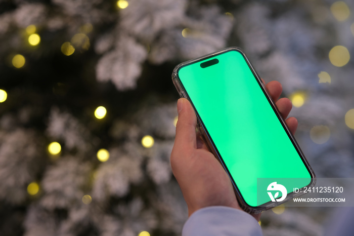 hand holding green screen smartphone in front of blur Christmas tree. Over the shoulder