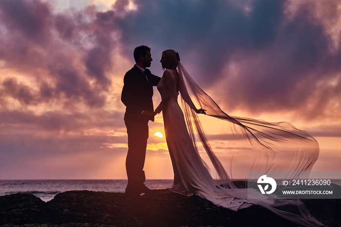 Wedding couple holds each other hands standing on the beach against the amazing sunset.