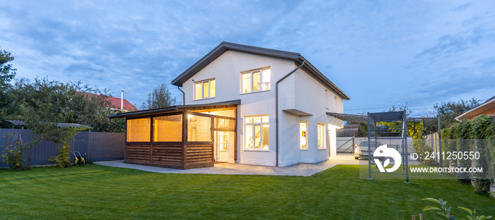 Exterior view of modern white house with courtyard, patio area, green grass lawn, garden and a car in the evening