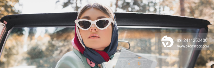 Elegant woman in sunglasses and headscarf holding map in roofless auto, banner