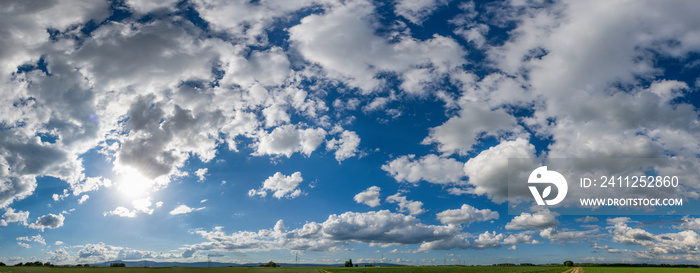 Panoramaansicht von Schönwetterwolken mit Horizont am unteren Bildrand und blauem Himmelim Gegenlicht der am Himmel stehenden Sonne