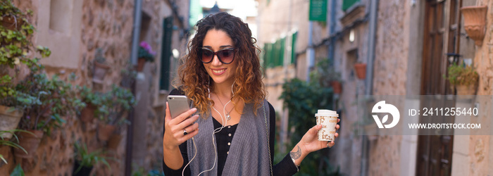 Young woman walking the cobblestone streets of an old tourist sea town while drinking coffee and listening music on earphones