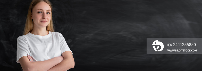 Teacher girl 20s millennial standing in front of the blackboard in the classroom. Blond student indoors near a black blank board. Place for the text on the right. Banner.