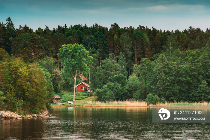 Sweden. Beautiful Red Swedish Wooden Log Cabin House On Rocky Island Coast In Summer. Lake Or River And Forest Landscape