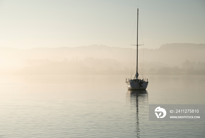 Stunning unplugged fine art landscape image of sailing yacht sitting still in calm lake water in Lake District during peaceful misty Autumn Fall sunrise