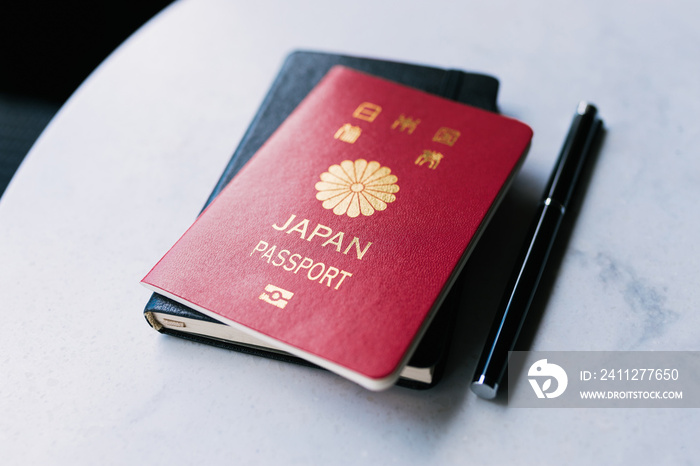 One single red Japanese passport laying on top of a black notebook next to a black pen that is on a white table