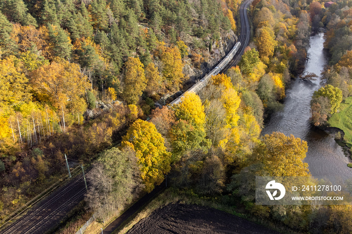 A train passing by in autumn landscape. Aerial, drone photography taken from above in Sweden in October. Trees, forest in vibrant colors and a river with flowing water.