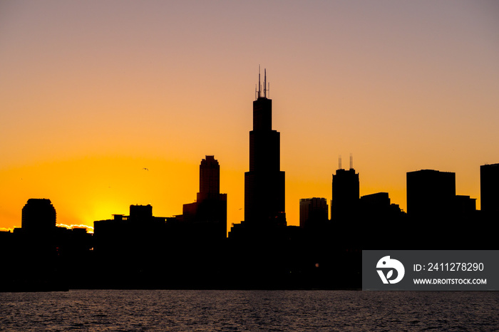 Chicago skyline picture during beautiful orange yellow sun as it lowers below the building silhouettes and the water of lake Michigan in the foreground