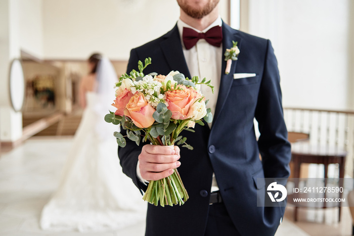 Close up of beautiful bridal bouquet of coral roses and greenery in groom’s hand with bride in blur on background indoors, copy space. Happy wedding couple before ceremony