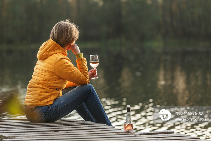 Woman in a yellow jacket relaxing on a wooden pier on a lake with a glass of rose wine watching fall sunset alone.