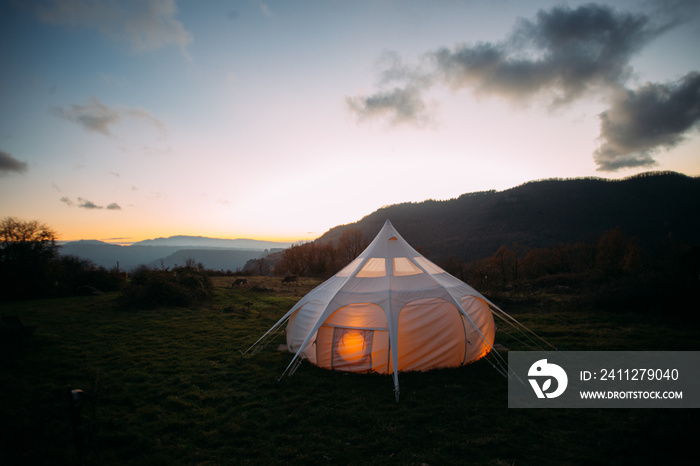 Night time sunset shot of glamping camping tent set up in beautiful nature landscape. Cosy and warm big tent lit from inside with orange dimmed lights. Romantic weekend getaway for couple