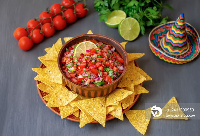 Pico de Gallo. Bowl of authentic chunky salsa made from tomatoes, onion, cilantro, chile peppers and lime juice served on a plate with tortilla chips. Dark background, selective focus, copy space.