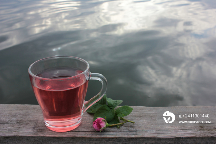 Glass cup of herbal tea with a pink tea rose petals on the wooden pier over the river. Summer tea party outdoors